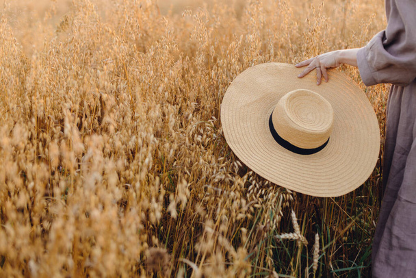 Stylová žena se slaměným kloboukem kráčející po ovesném poli ve světle zapadajícího slunce, s oříznutým výhledem. Mladá fena v rustikálních lněných šatech odpočívá ve večerní letní krajině. pomalý život na venkově - Fotografie, Obrázek