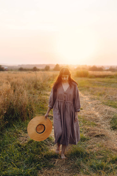 Mulher elegante com chapéu de palha andando no campo de aveia em luz do pôr do sol. Momento de tranquilidade atmosférica. Jovem fêmea em vestido de linho relaxante no verão à noite campo, rural vida lenta - Foto, Imagem