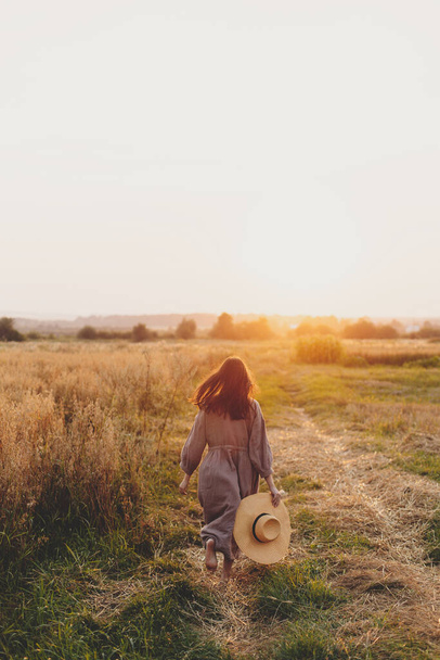 Стильна жінка з солом'яним капелюхом біжить на вівсяному полі під час заходу сонця. Атмосферний щасливий момент. Молода жінка в сільській лляній сукні відпочиває в вечірній літній сільській місцевості, сільському житті
 - Фото, зображення