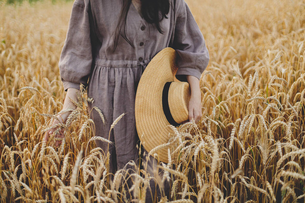 Femme avec chapeau de paille tenant des tiges de blé en plein champ, vue dégagée. Moment de tranquillité atmosphérique. Femme en robe de lin rustique touchant les épis de blé mûr dans la campagne estivale. Vie rurale lente - Photo, image