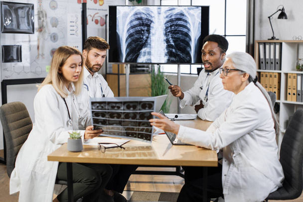 African american male doctor showing his colleagues pulmones disease on digital display screen. Team of multiethnic physicians sitting at room and discussing various way of treatment. - Foto, Imagem