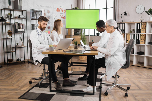 Team of multicultural medical specialists with thoughtful facial expression looking on monitor with green screen. Male and female doctors in lab coats having regular sessions in medical sphere. - Фото, изображение
