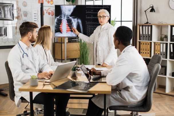 Qualified female doctor leading seminar to three multiracial interns at boardroom. Senior woman standing near digital screen and explaining results of chest x ray scan. - Foto, imagen