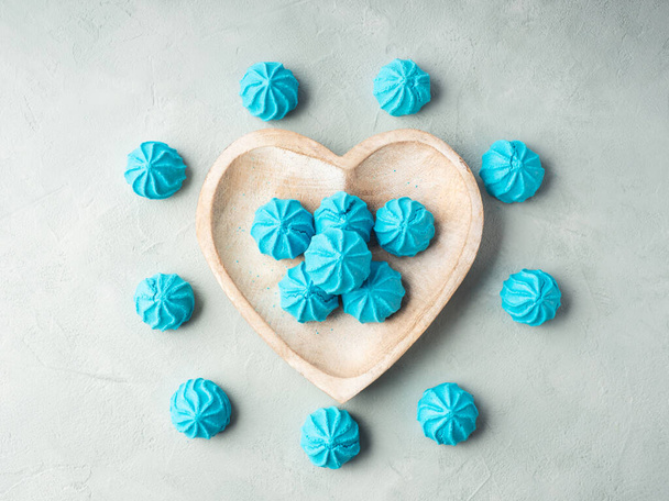 Plato de madera en forma de corazón con merengues dulces azules sobre fondo de piedra gris. Puesta plana - Foto, imagen