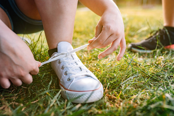 close-up of the hands of a young girl bending down, tying her white trainers in a public park in the city. concept of leisure and free time. outdoor, natural sunlight, grass in background. - Fotó, kép