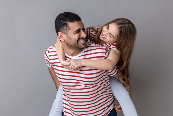 Joyful father giving piggyback ride for his charming daughter, smiling girl looking at her daddy with love, having fun and playing together. Indoor studio shot isolated on gray background. - Zdjęcie, obraz