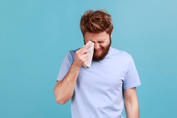 Portrait of unhappy frustrated bearded man sobs and rubs tears with handkerchief feels desperate, has dissatisfied and sorrow expression. Indoor studio shot isolated on blue background. - Foto, imagen