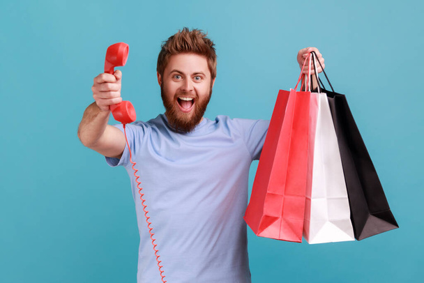 Portrait of excited positive bearded man holding shopping bag and red retro telephone, call me, looking at camera with excitement. Indoor studio shot isolated on blue background. - Foto, Imagen