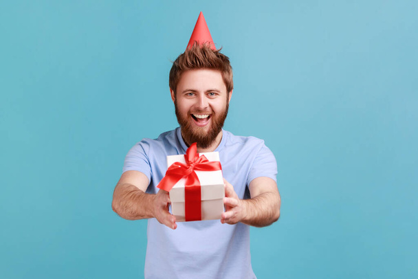 Portrait of man in party cone giving birthday gift and smiling, sharing present, donating and offering surprise in box, congratulation on anniversary. Indoor studio shot isolated on blue background. - Foto, Bild