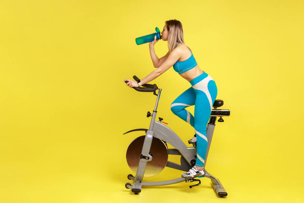 Side view portrait of blonde woman training on exercise bike and drinking water, refreshing, replenishes water balance, wearing blue sportswear. Indoor studio shot isolated on yellow background. - Foto, afbeelding