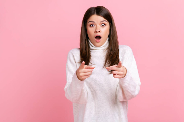 Extremely astonished female pointing at camera, being very shocked to choose you, keeps mouth open, wearing white casual style sweater. Indoor studio shot isolated on pink background. - Foto, afbeelding