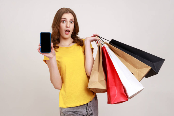 Awesome online purchase! Portrait of amazed teen girl holding cell phone and shopping bags, advertising delivery apps, looking at camera with open mouth. Indoor studio shot isolated on gray background - Фото, изображение