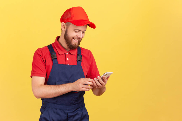 Portrait of satisfied smiling courier or craftsman standing with with smartphone in hands, taking online order, being ready to work. Indoor studio shot isolated on yellow background. - Photo, image