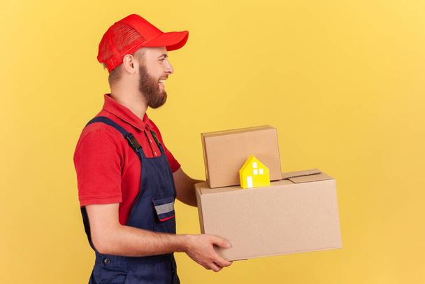Profile portrait of delivery man in uniform and red cap holding cardboard boxes and paper house, door-to-door delivery, help in relocating. Indoor studio shot isolated on yellow background. - Foto, Imagen