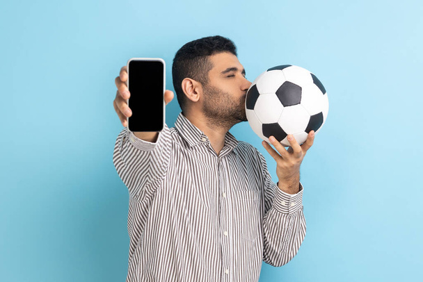 Portrait of bearded handsome man standing kissing soccer ball and showing mobile phone with empty black display, wearing striped shirt. Indoor studio shot isolated on blue background. - Fotografie, Obrázek
