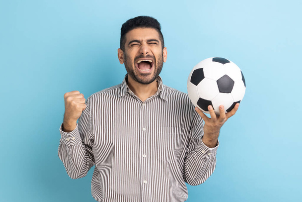 Bearded businessman screaming widely opening mouth, celebrating victory of favourite football team, holding soccer ball in hands, wearing striped shirt. Indoor studio shot isolated on blue background. - Fotoğraf, Görsel