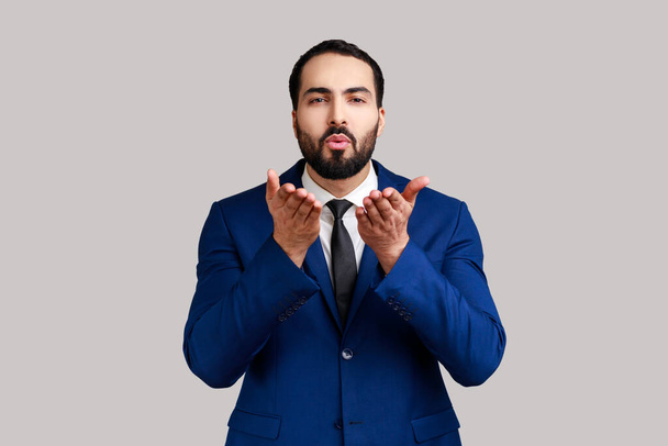 Handsome bearded man sending air kiss over palms, flirting to camera, demonstrating romantic amorous feelings, wearing official style suit. Indoor studio shot isolated on gray background. - Foto, Bild