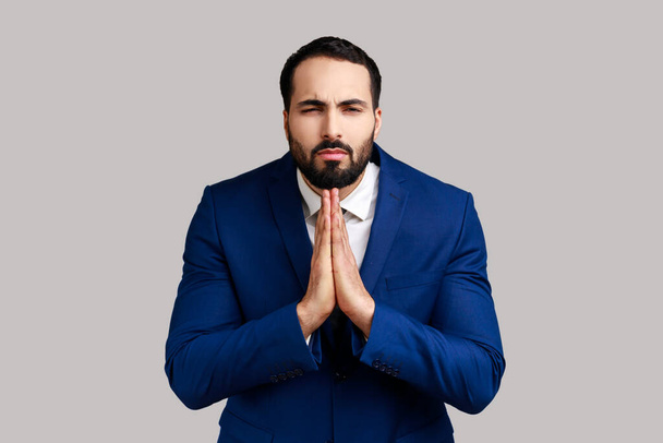 Portrait of man holding hands in prayer, looking with imploring pleading expression, begging help, asking forgiveness, wearing official style suit. Indoor studio shot isolated on gray background. - 写真・画像