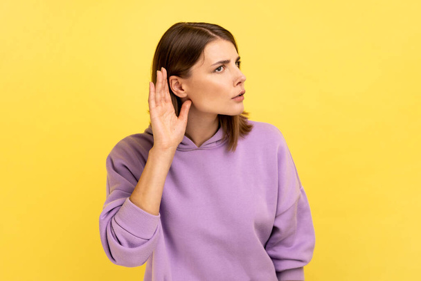 Portrait of pretty woman holding hand near ear, listening attentively with interest private conversation, confidential talk, wearing purple hoodie. Indoor studio shot isolated on yellow background. - Foto, Imagem