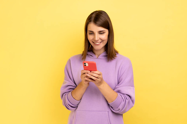 Portrait of happy pretty woman reading message on smartphone and smiling, using mobile device for communication, browsing web, wearing purple hoodie. Indoor studio shot isolated on yellow background. - Fotografie, Obrázek