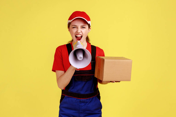 Portrait of angry courier woman holding cardboard box and screaming in megaphone, looking at camera with aggressive expression, wearing overalls. Indoor studio shot isolated on yellow background. - Foto, immagini