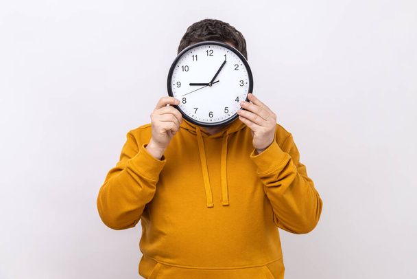 Portrait of anonymous unknown man hiding face behind big wall clock, time management, reminding of deadline, wearing urban style hoodie. Indoor studio shot isolated on white background. - Zdjęcie, obraz