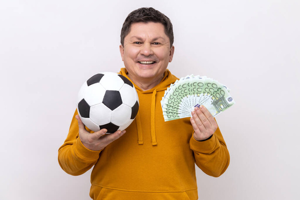 Gülümseyen orta yaşlı bir adam futbol topu gösteriyor ve 100 Euro 'luk banknotlarla eğleniyor spor bahislerinde çok para kazanıyor, şehir tarzı kapüşonlu kapüşonlu giyiyor. Beyaz arka planda izole edilmiş kapalı stüdyo görüntüleri. - Fotoğraf, Görsel