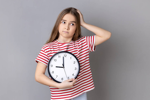 Portrait of confused uncertain little girl wearing striped T-shirt holding big wall clock in hands, looking away with puzzled expression. Indoor studio shot isolated on gray background. - Fotografie, Obrázek