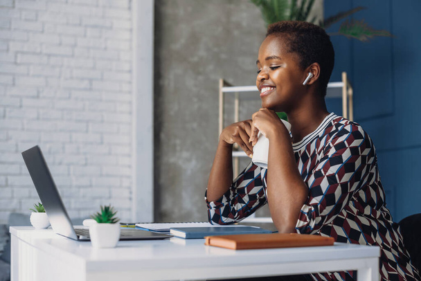 Glimlachend Afrikaans-Amerikaanse vrouwelijke freelancer die aan het bureau zit, een online cursus volgt, naar een laptop scherm kijkt. Internettechnologie. Ondernemingen - Foto, afbeelding
