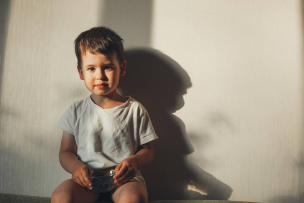 Παιδί που κρατά ένα μπολ με βατόμουρα βιολογικό προϊόν, κάθεται στον καναπέ στο σπίτι. - Φωτογραφία, εικόνα