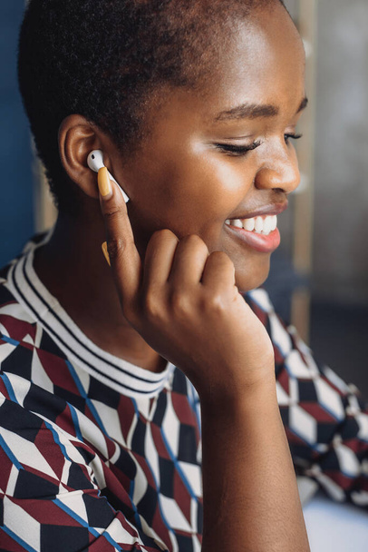 Close-up portrait of an African woman holding her hand up to her wireless headset and smiling as she listens to her favorite music. Businesswoman portrait - Foto, Imagem