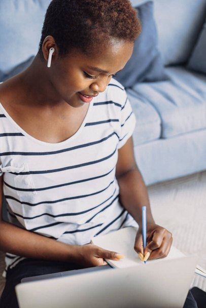 Musta nainen työskentelee kaukana kotoa opiskelemassa koulun oppituntia olohuoneessa istuessaan lattialla. Amerikkalainen musta. Tietotekniikan selittäminen - Valokuva, kuva