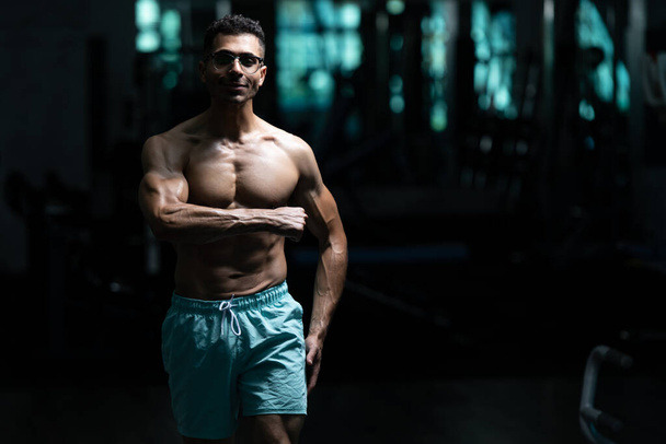 Όμορφος νεαρός άνδρας με γυναικομαστία Στήθη Πρόβλημα στέκεται ισχυρή στο γυμναστήριο και κάμψη των μυών - Φωτογραφία, εικόνα