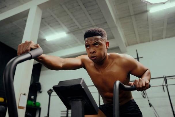 African American male using an elliptical trainer during cross fit training. Male personal trainer exercising intensely in the gym. High quality photo - Фото, изображение