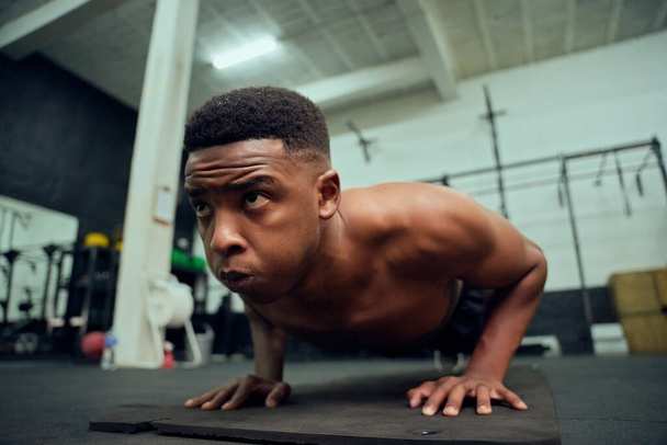 Motivated African American male doing push-ups in the gym. Mixed race, male personal trainer doing cross fit indoors. High quality photo - Photo, Image