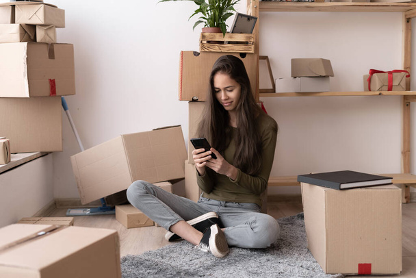 Yeni evin oturma odasında akıllı telefon kullanan mutlu, genç bir İspanyol kadın taşınma gününde bir yığın karton kutuyla, gülümsüyor. - Fotoğraf, Görsel