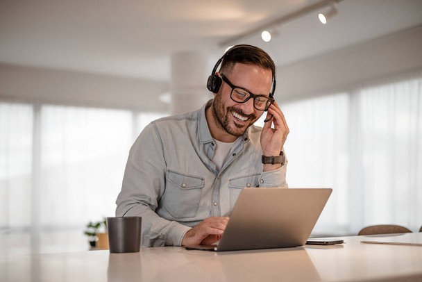 Young smiling businessman video conferencing on laptop. Happy professional discussing through headset at desk. He is working in corporate office. - Valokuva, kuva