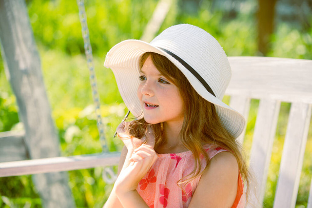 A gyerek hintázik. Egészséges nyári tevékenység gyermekek számára meleg időben. A kisgyerekek hintáznak. Közel portré egy gyönyörű lány kalap álmodik. Álmodozó gyerekek arca - Fotó, kép