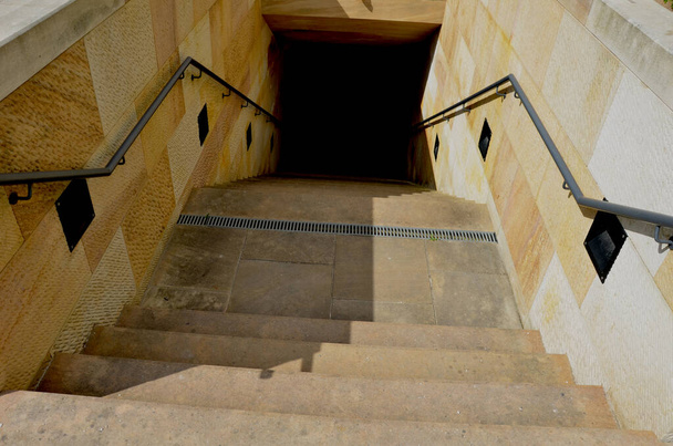 Die beige Treppe führt unter das Gelände eines Rasenhofes mit Parkwegen hinab. Die Wände bestehen aus bearbeiteten Sandsteinblöcken. Hintergrundbeleuchtung Treppen mit Halogenen an den Seiten - Foto, Bild