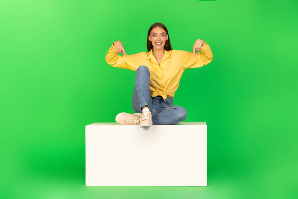 Nagyszerű ajánlat. Izgatott nő ujjal lefelé ülő fehér stand reklám valami pózoló mosolygós kamera felett zöld háttér stúdió. Nézd, ott koncepció. - Fotó, kép