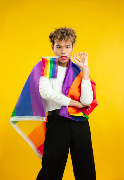 young gay with pride movement LGBT Rainbow flag on shoulder isolated yellow background. Man with a gay pride flag looking at camera. - Valokuva, kuva