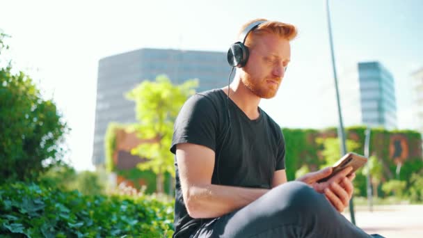 Confident red haired man listening music in headphones and typing by phone while sitting on the bench outdoors - Felvétel, videó