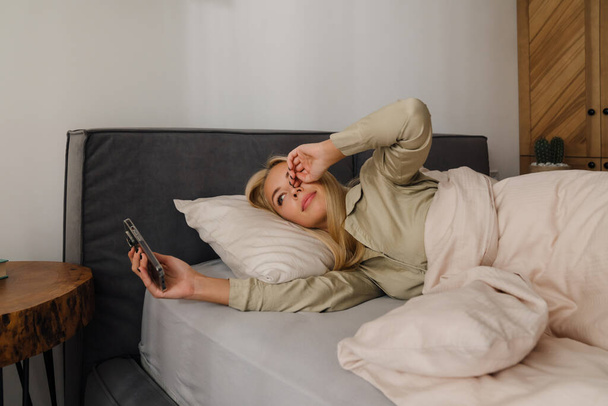 Young blonde woman using mobile phone while lying in bed after sleep at home - 写真・画像