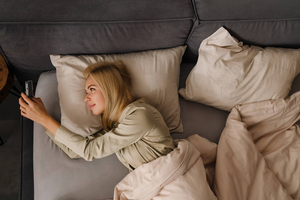 Fiatal szőke nő használ mobiltelefon, miközben feküdt az ágyban alvás után otthon - Fotó, kép