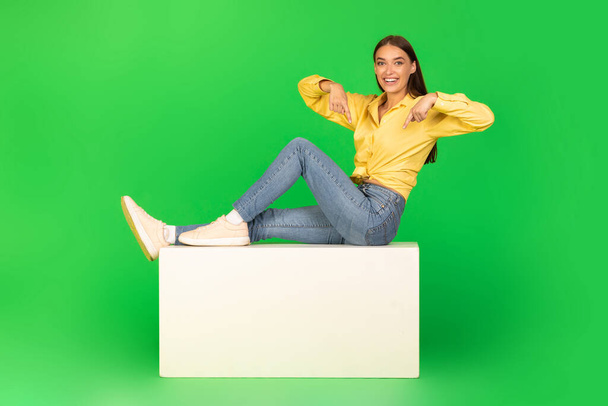 Şuraya bak. Yeşil Stüdyo 'nun arka planında oturan kameraya gülümseyen Mutlu Kadın Parmaklarını Beyaz Stantta Gösteriyor. Harika Teklif, Metin Reklam Kavramı - Fotoğraf, Görsel