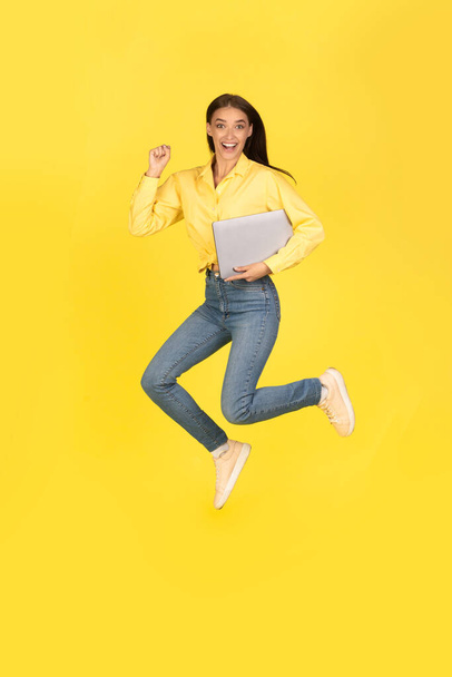 Joyful Lady saltando sosteniendo la computadora portátil agitando puños en alegría celebrando la suerte sonriendo a la cámara posando en medio del aire sobre el fondo amarillo del estudio. Internet y Tecnología. Vertical - Foto, imagen