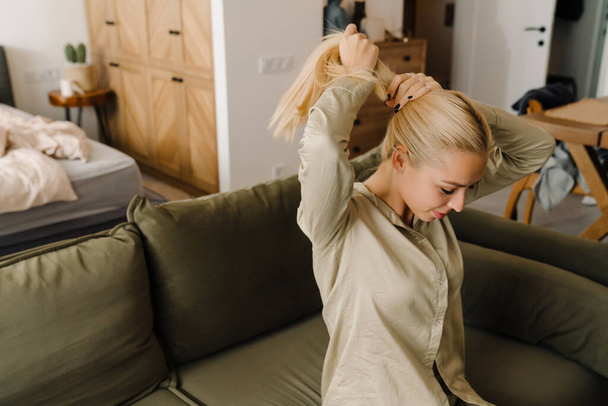 Blonde white woman doing hairstyle while sitting on sofa at home - Φωτογραφία, εικόνα