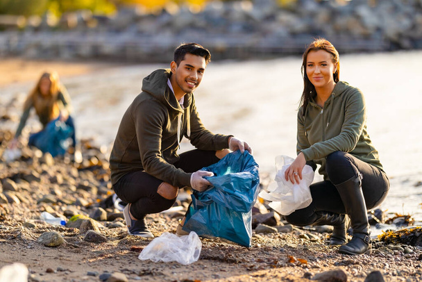 Equipo de voluntarios dedicados y sonrientes recogiendo basura de plástico en la bolsa en el día soleado. Hombres y mujeres están limpiando la playa rocosa. Están protegiendo el ecosistema. - Foto, imagen