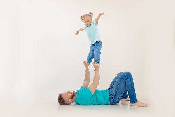 Athletic man with pumped up arms lying on back holding and lifting up with hands standing little girl, white background. Whole family workout, father and daughter. Sport and healthy lifestyle concept. - Fotografie, Obrázek