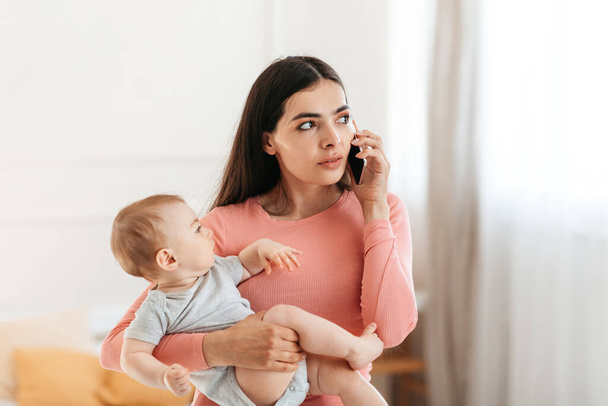 Lebensstil junger Mütter. Nachdenkliche Mutter, die mit dem Handy telefoniert und Baby im Arm hält, Arzt anruft oder zu Hause telefoniert, freier Raum - Foto, Bild